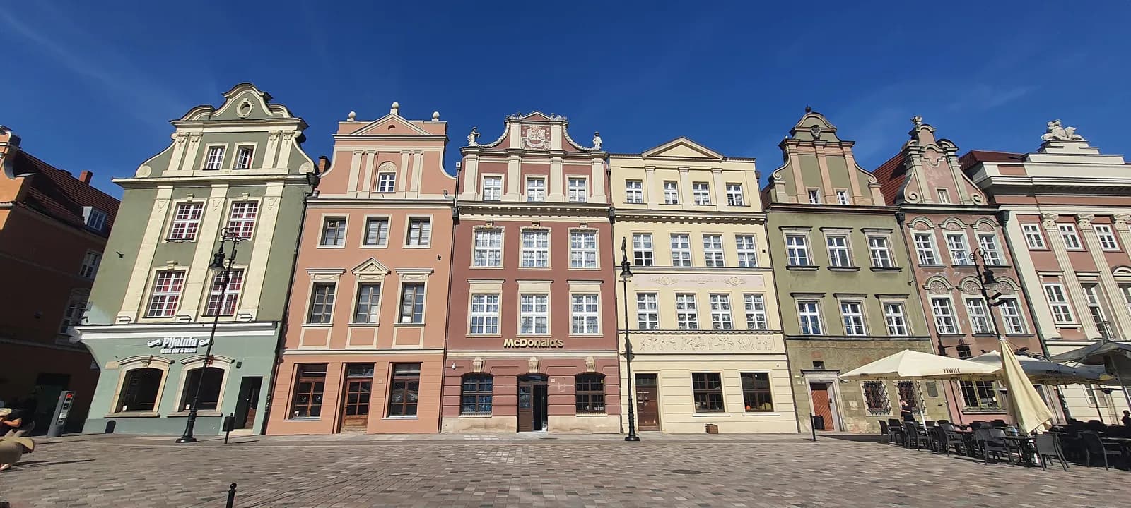 A row of colorful, historic buildings with gabled roofs and numerous windows lines a cobblestone square under a clear blue sky, with outdoor cafe seating visible on the right.