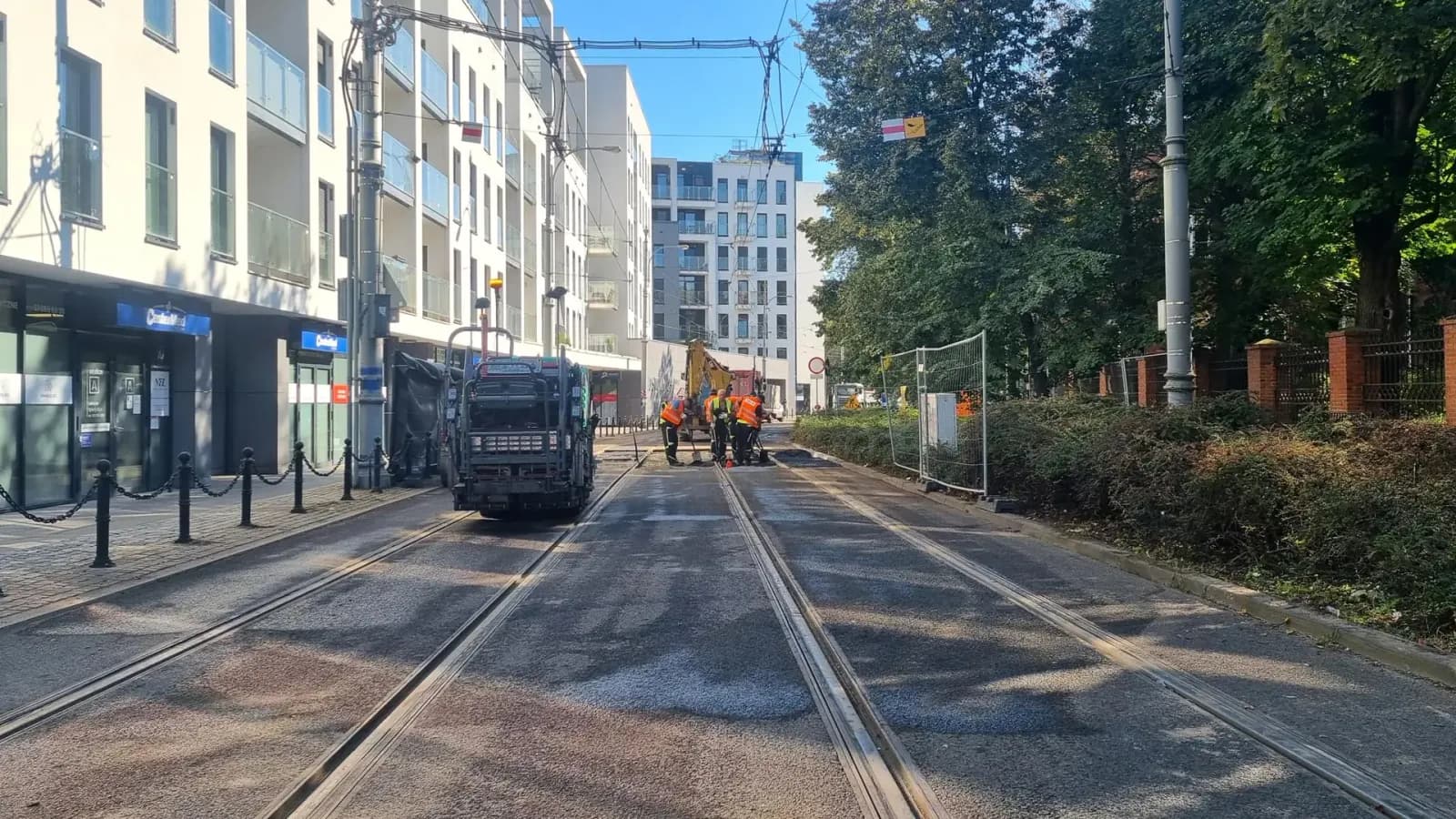 Construction workers in orange vests working on tram tracks in the middle of a street, with residential buildings and trees flanking both sides.