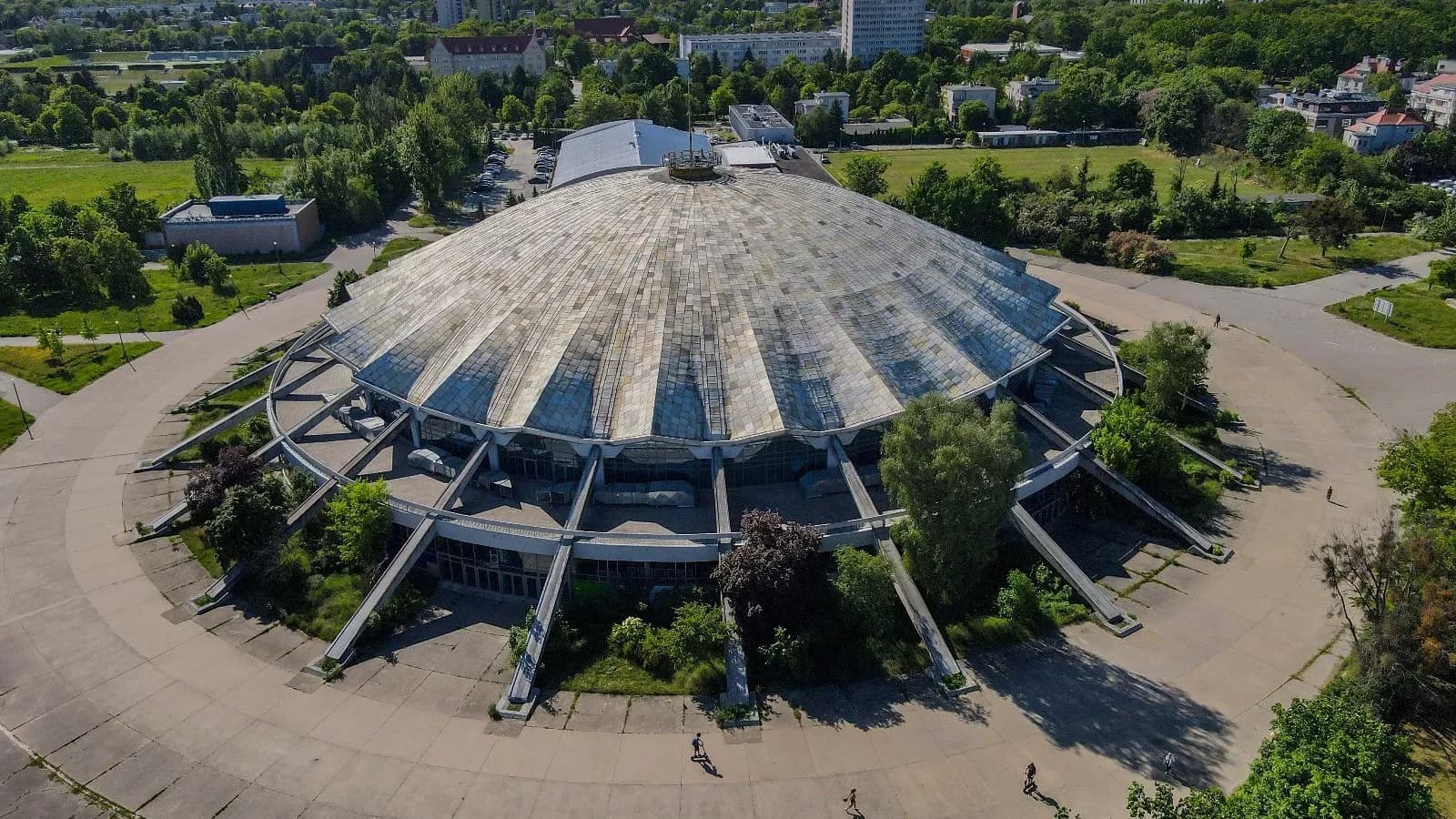 An aerial view shows a large, round, domed building with a ribbed roof, surrounded by green trees and asphalt paths, with other buildings and more greenery in the background.