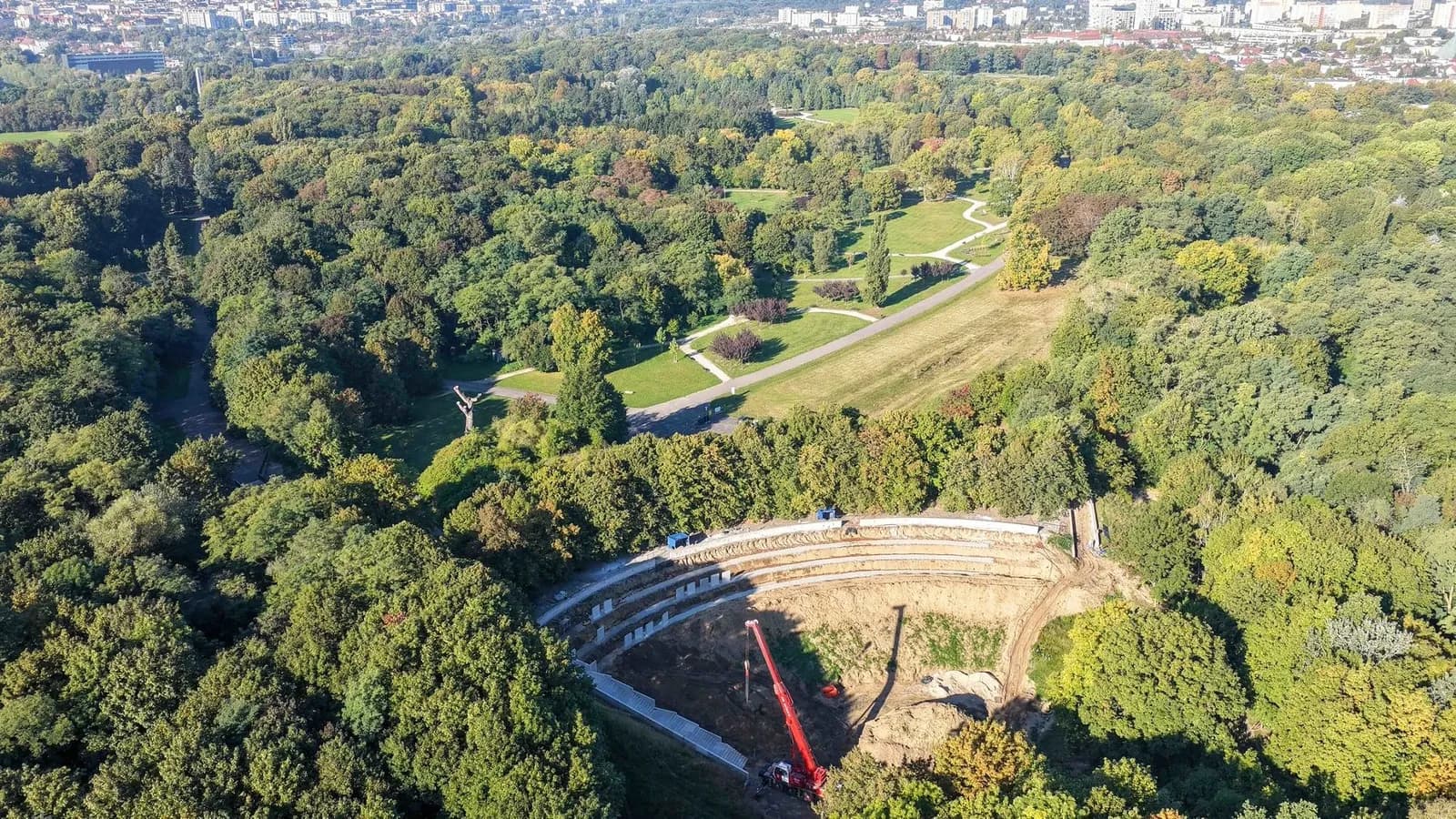 An aerial view of an amphitheater under construction within a lush green forest, with a red crane and nearby city buildings in the distance.