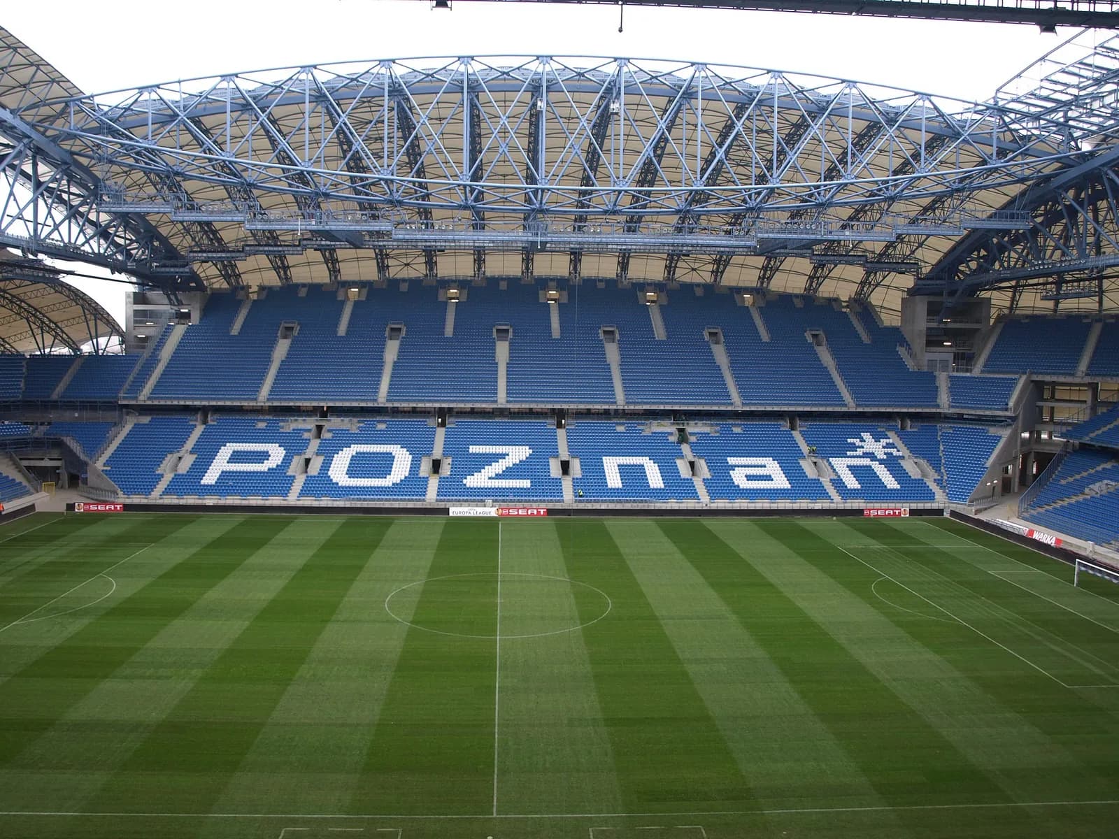 An empty football stadium with green grass field and blue seats, where the word "POZNAN" is spelled out in white letters on the seats.