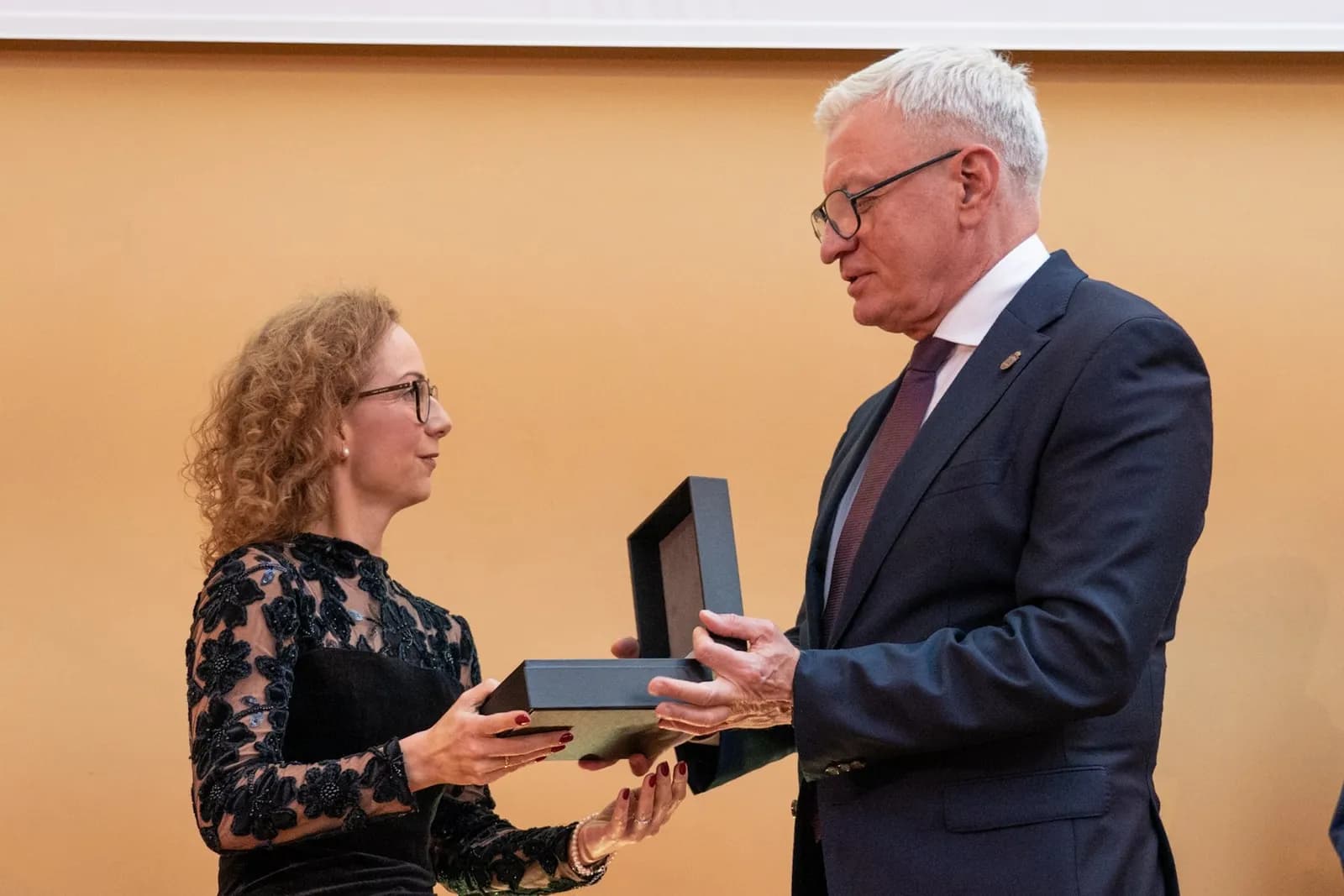 A man in a suit presents an open black box to a woman in a black dress with lace sleeves.