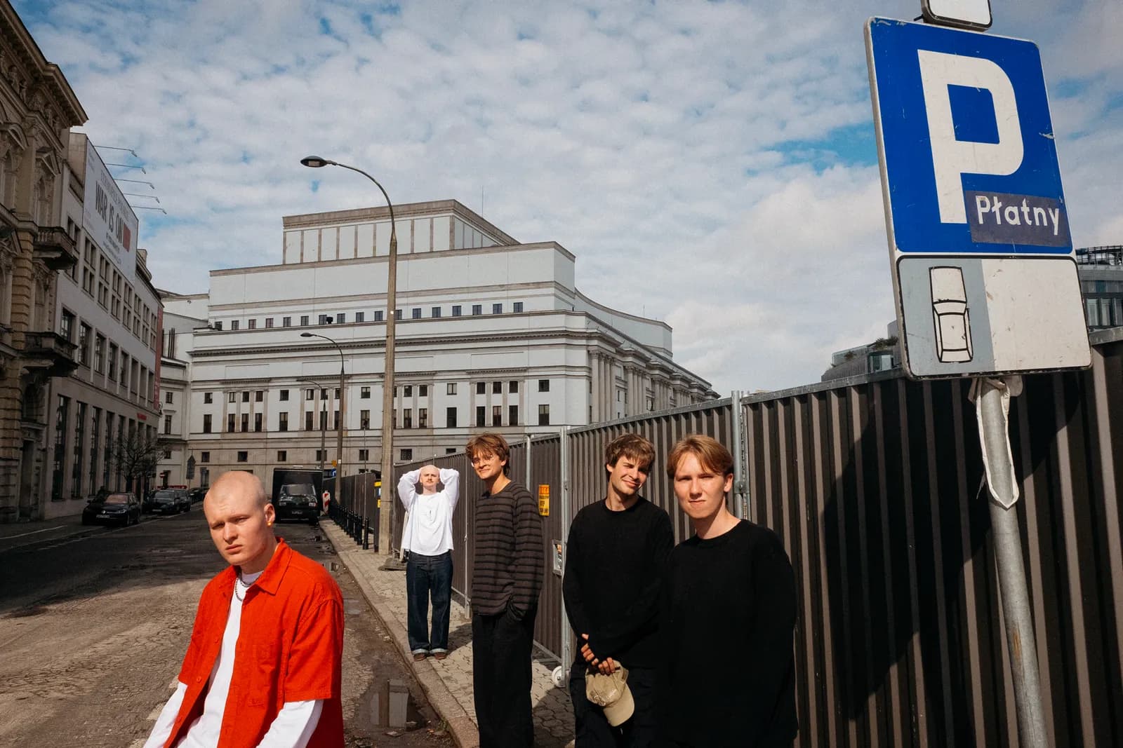 Four young men stand on a city street, with one man in a red shirt in the foreground, and a large white building in the background beneath a blue sky.