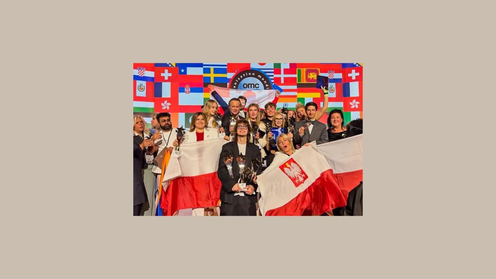A large group of people hold up trophies and Polish flags in front of a backdrop of international flags.