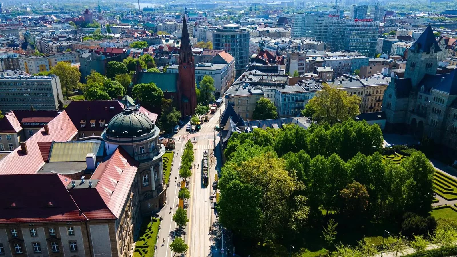 An aerial view of a European city with a mix of old and new architecture, including a prominent red-brick church spire, a large domed building, and a street with tram lines lined with trees.