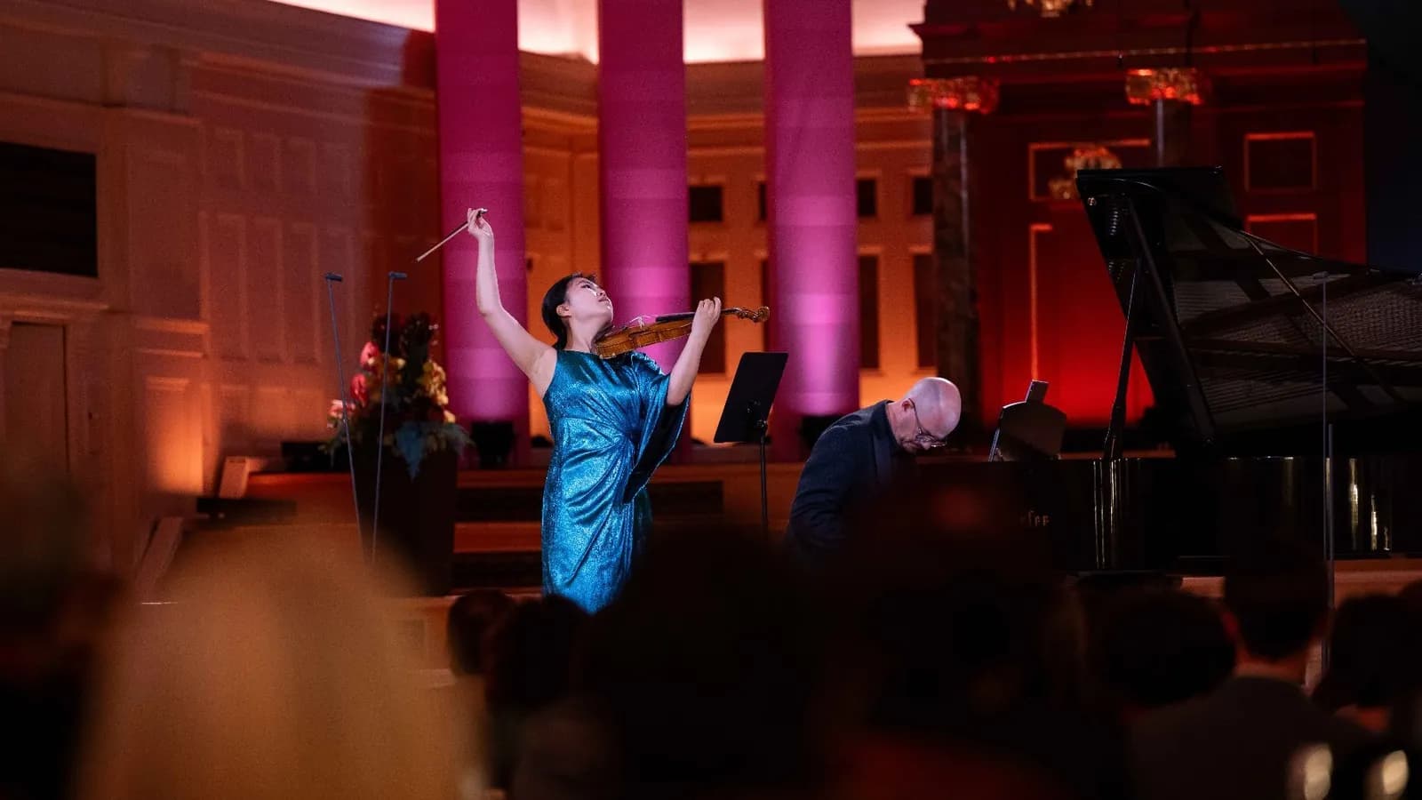 A woman in a shimmering blue dress plays the violin on stage, her arm raised with the bow, while a bald man plays the piano beside her.