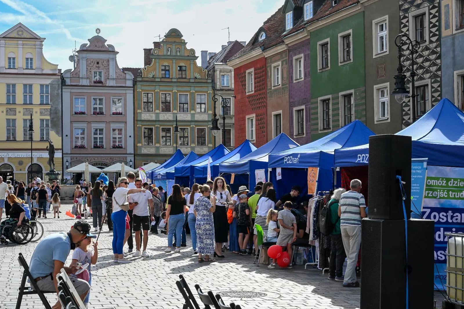 A lively outdoor market scene with people browsing blue tents, set against a backdrop of colorful historic buildings under a sunny sky.