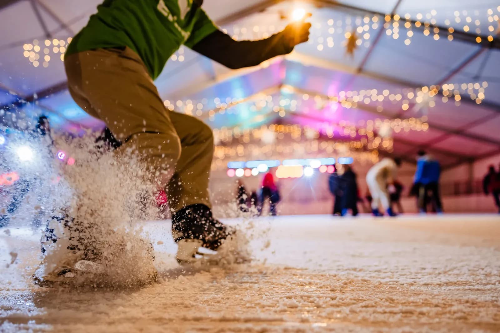 A person wearing ice skates and brown pants kicks up a spray of ice on an indoor rink, with blurred figures and string lights in the background.