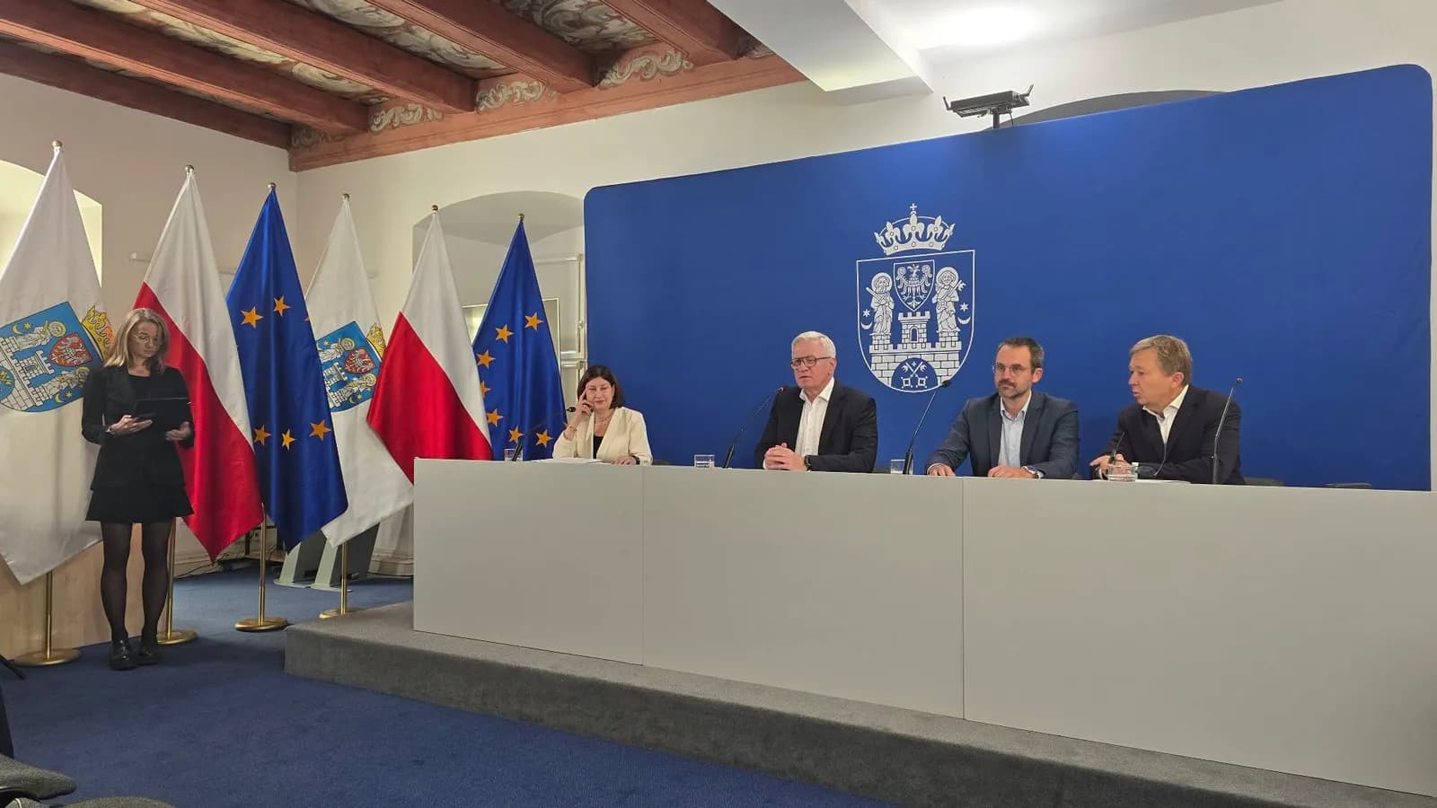 A woman stands to the left of four men sitting behind a long table, all in front of a blue backdrop with a coat of arms and several Polish and European Union flags.