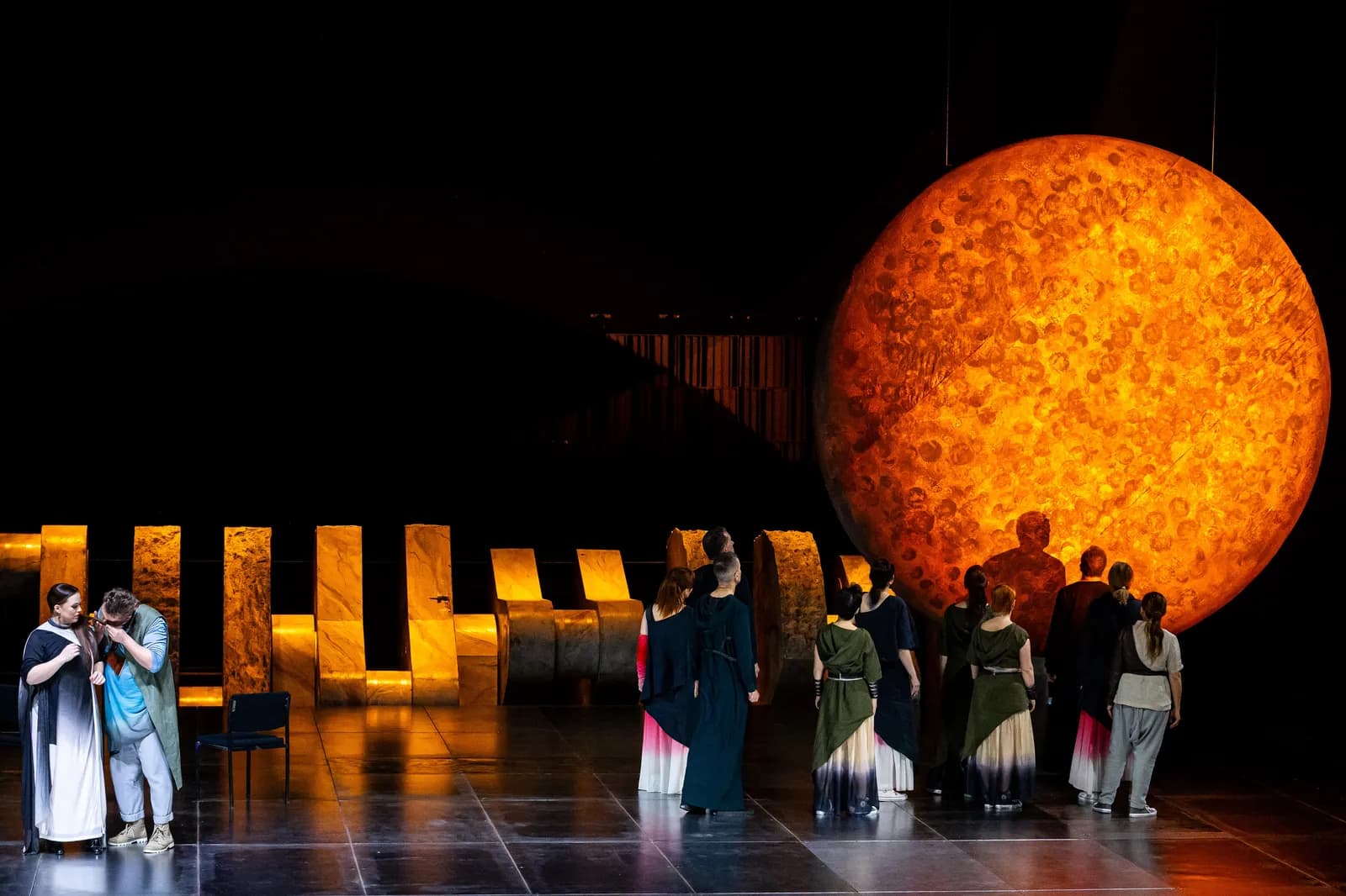 Performers on stage during an opera with a large, glowing orange orb resembling the moon or sun in the background, as a group of people stand facing it.