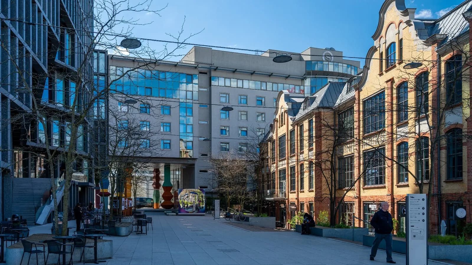 A wide outdoor plaza with modern glass buildings on the left and traditional brick buildings with ornate gables on the right, under a clear blue sky, with a few people walking.