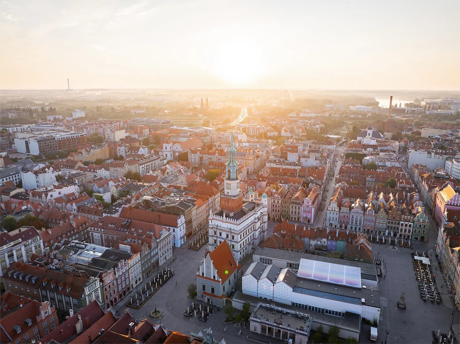 An aerial view of a European city at sunset, featuring a prominent historic town hall with a tall spire surrounded by colorful buildings and a bustling market square.