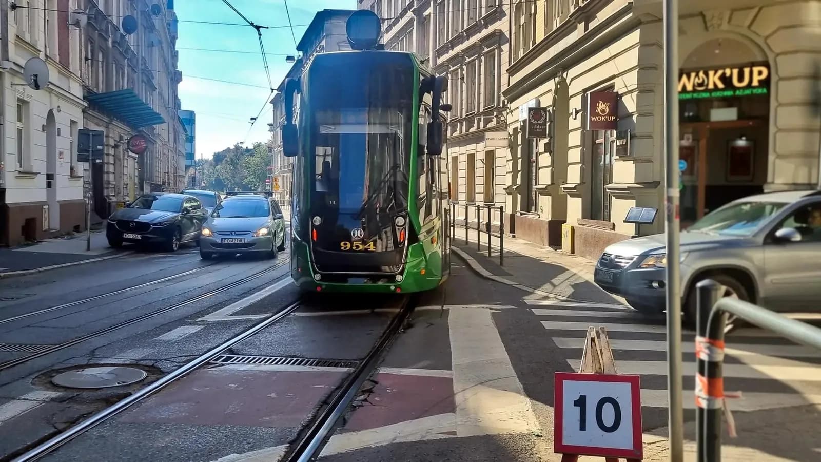 A green and black tram numbered 954 travels down a city street with cars parked and driving alongside it under a bright sky.