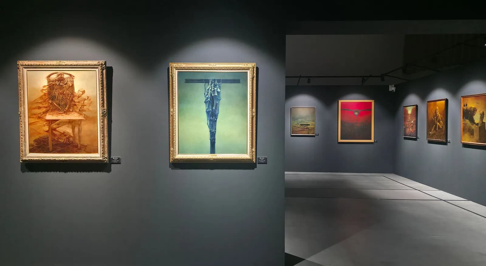 A dimly lit art gallery showcases several framed paintings with dark and surreal themes on a gray wall, including two prominent works on the left and a series of smaller pieces extending into the background on the right.
