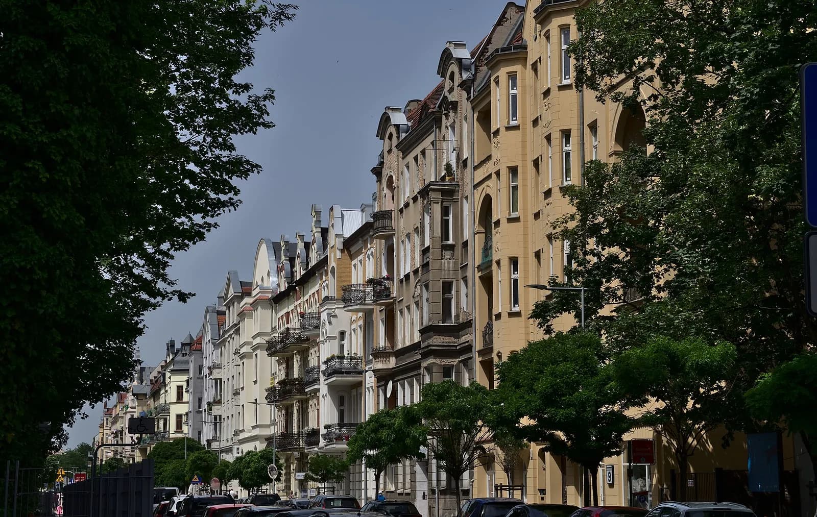 A long line of ornate, multi-story buildings with balconies and varied architectural styles, some light and others yellow-brown, line a street with trees and parked cars under a blue sky.