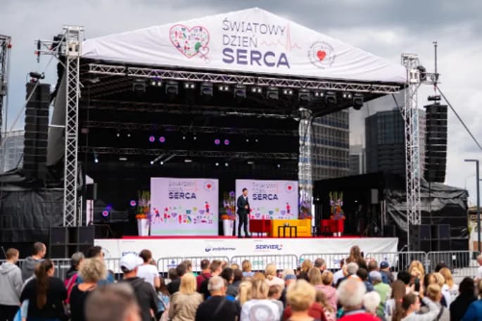 A man stands on a stage with two large screens behind him, addressing a crowd of people at an outdoor event.