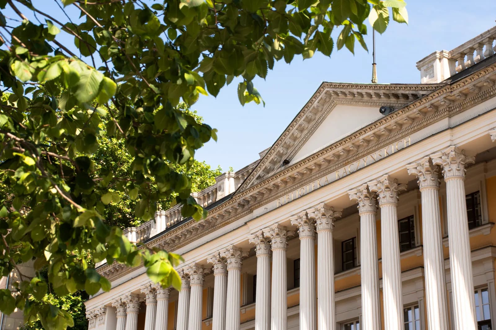 A grand white building with numerous classical columns and an ornate pediment, partially obscured by green leaves from a tree in the foreground.