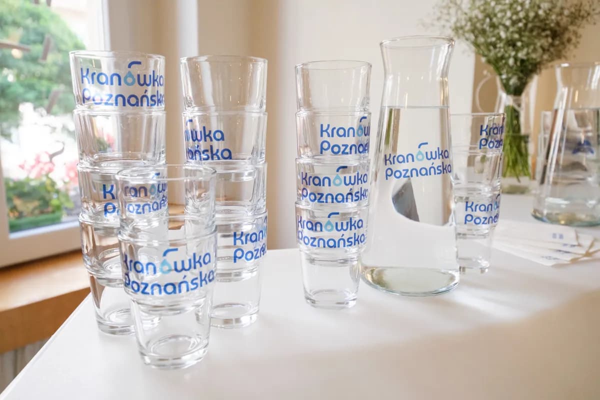 Multiple stacks of clear glasses and a carafe, all branded with blue text "Kranówka Poznańska," sit on a white table next to a window with greenery outside.