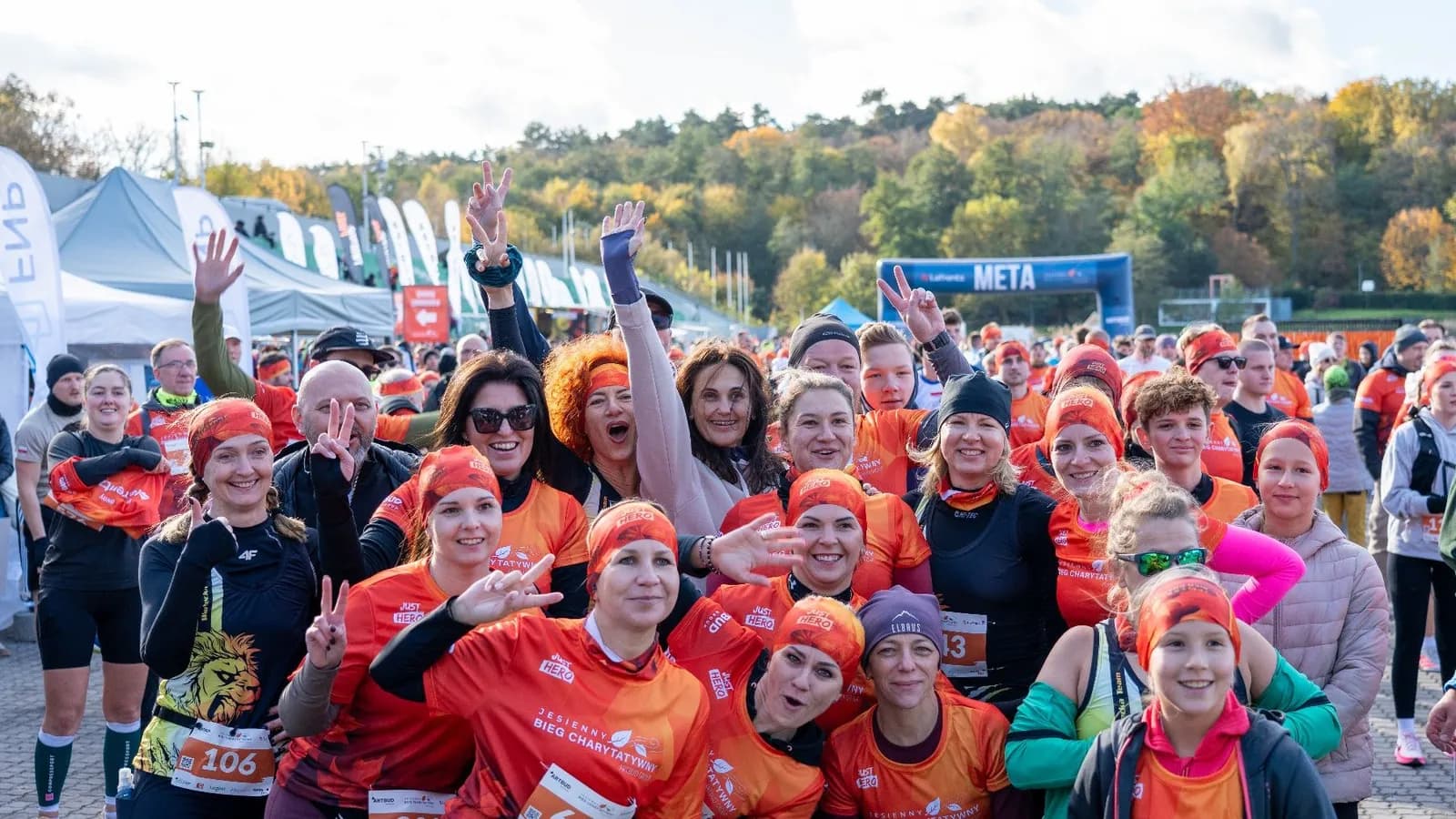 A large group of people in orange and black athletic wear posing for a photo with smiles and raised hands, some making peace signs, at what appears to be a charity run event with tents and trees in the background.