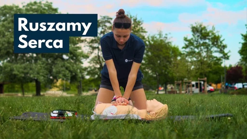 A person in a dark shirt and shorts performs chest compressions on a CPR manikin lying on a mat in a grassy park, with text overlayed on the top left.