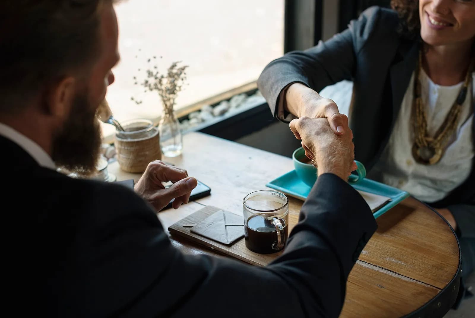 Two people in business attire shake hands across a wooden table with coffee cups and a phone, symbolizing a successful agreement or partnership.