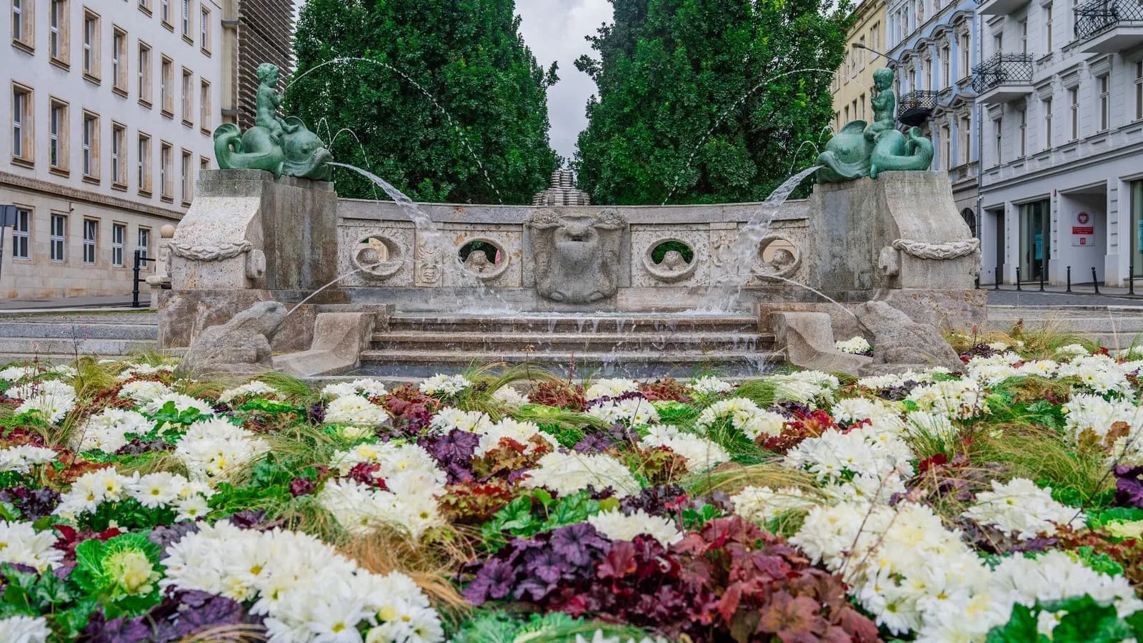 An ornate stone fountain with water flowing into tiered basins is surrounded by a vibrant flower bed with white, green, red, and purple plants, with buildings and trees in the background.