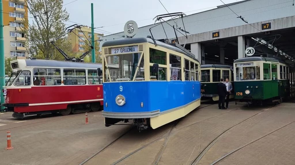 A line of vintage trams, red, blue, and green, parked in front of a tram depot with two men standing near the green tram.