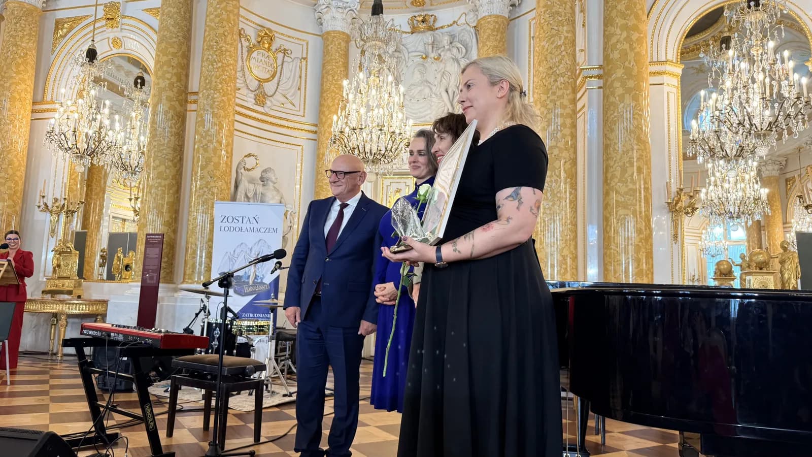 A woman in a black dress holds an award and a white rose while standing next to a man in a suit and another woman in a blue dress in a grand hall with chandeliers.