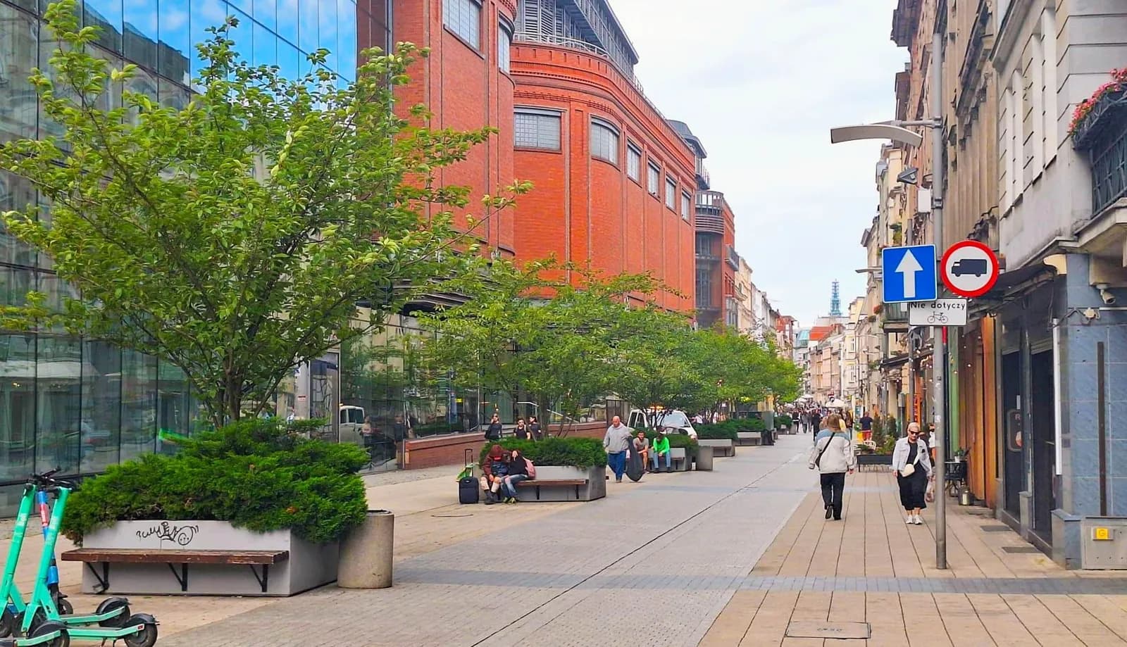 A wide pedestrian street lined with trees and benches, with people walking and sitting, and buildings on either side under a bright sky.
