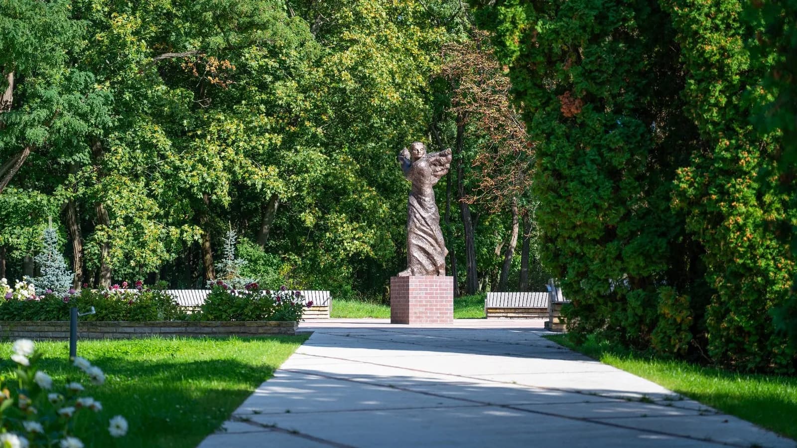 A bronze statue of a winged figure on a brick pedestal stands at the end of a paved pathway in a park, surrounded by lush green trees and benches.