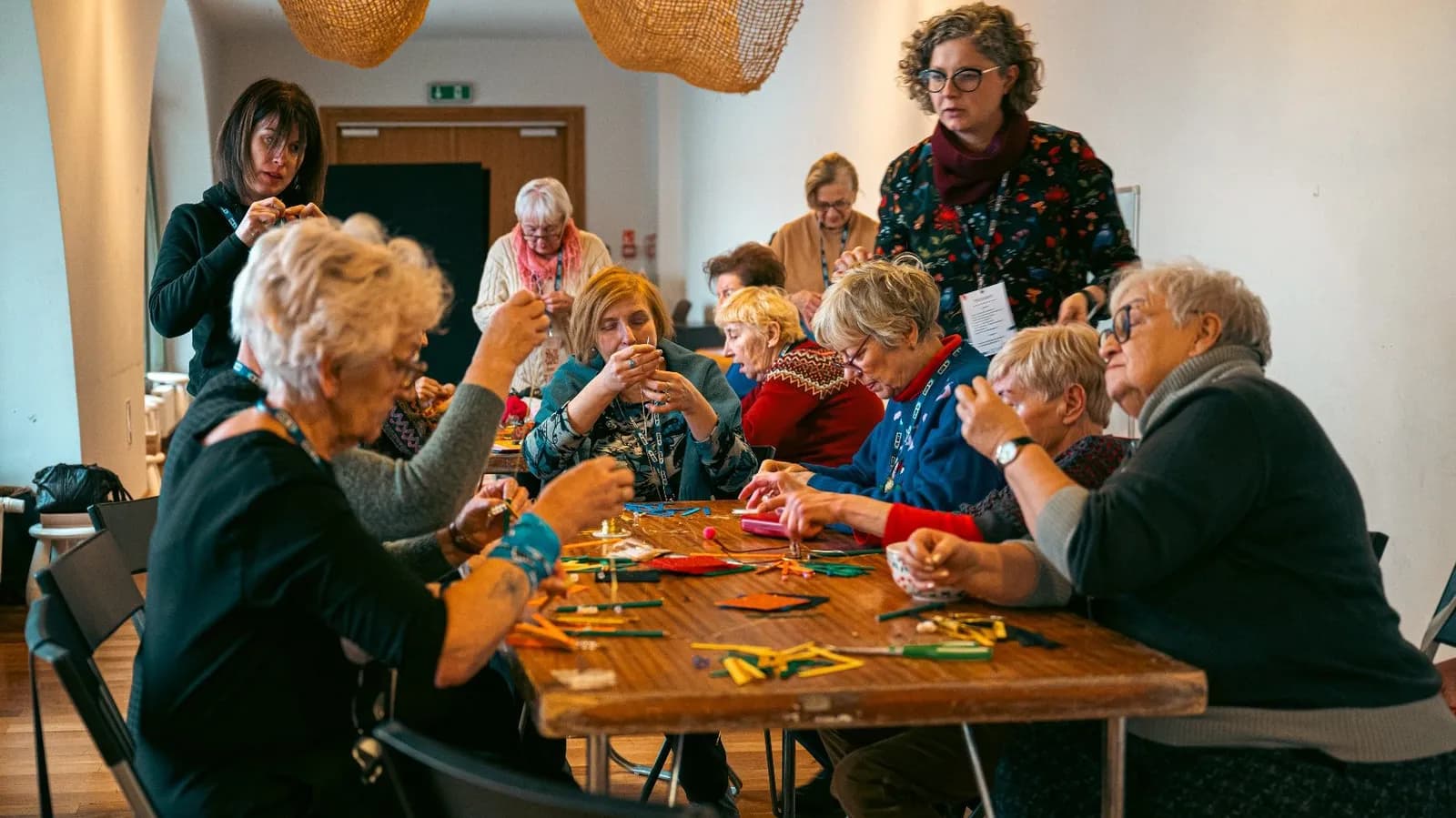 Several women are gathered around a wooden table, engaged in a craft activity, with one woman standing in the background seemingly observing or guiding.