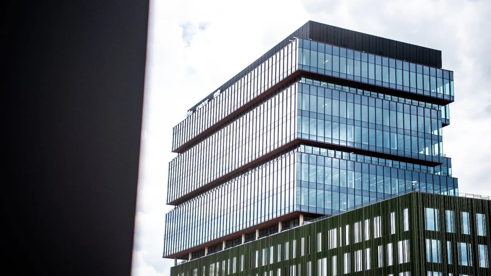 A modern building with a dark green base and upper floors composed of reflective blue glass, against a cloudy sky.