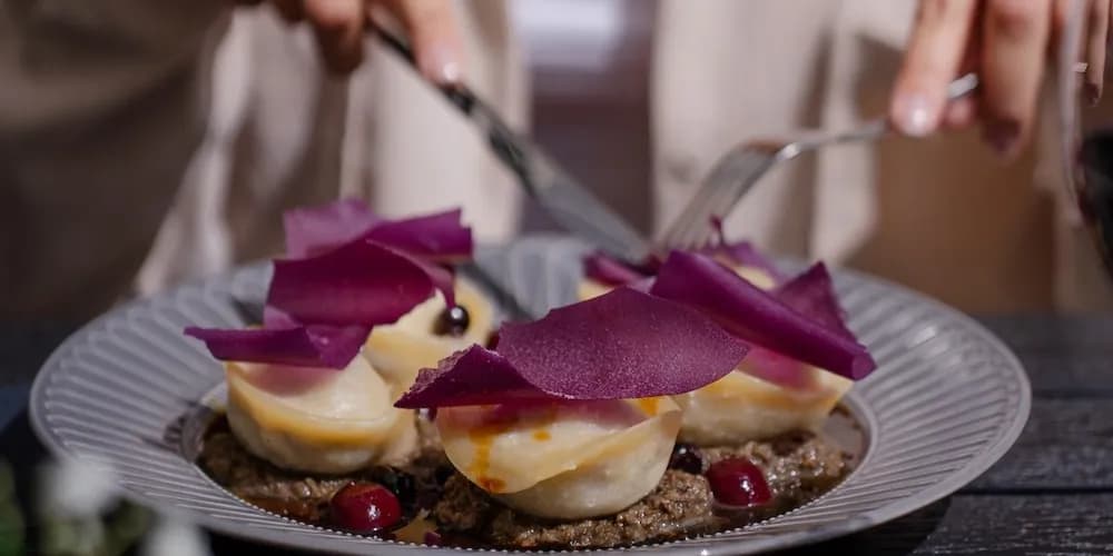 A person uses a fork and knife to cut into a beautifully plated dish featuring several small pastries topped with purple edible decorations, served on a bed of dark, textured sauce with red berries.