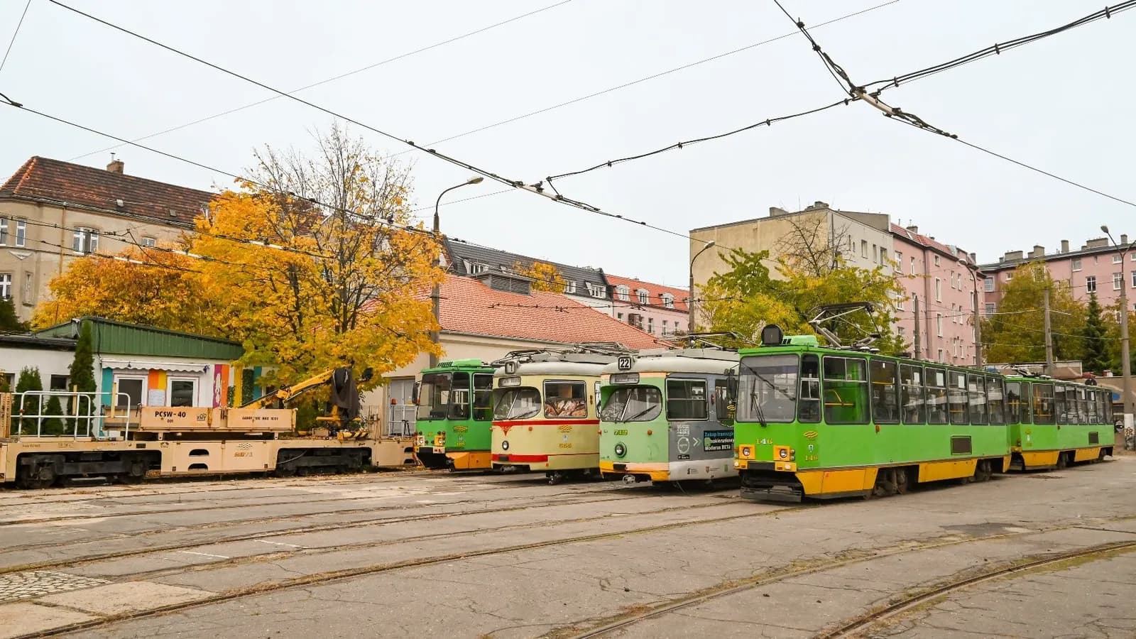 A line of vintage trams in various colors, including green, yellow, and cream, parked on tracks with buildings and autumnal trees in the background under an overcast sky.