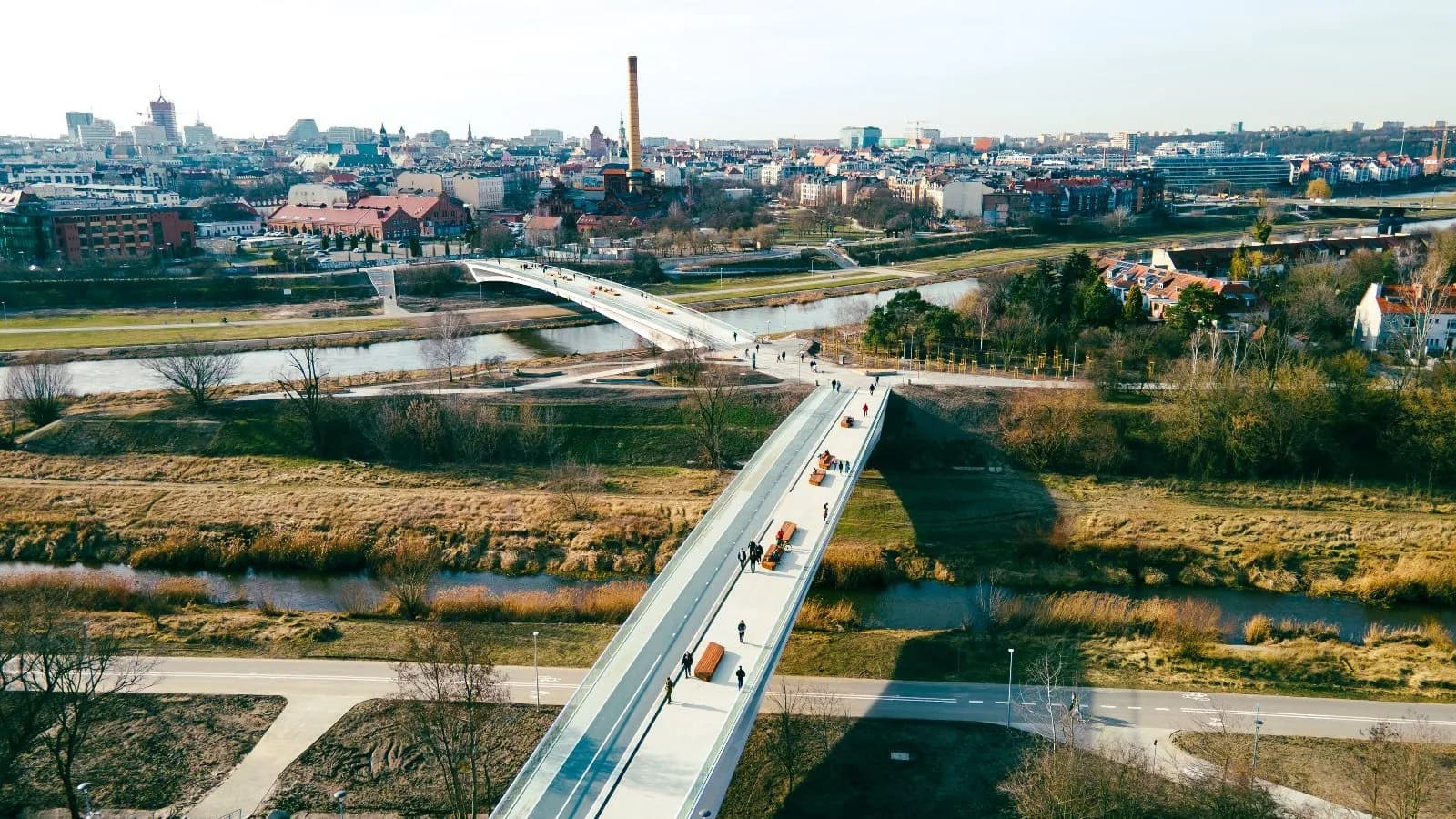 An aerial view of two modern pedestrian bridges spanning a river and a grassy park, leading towards a cityscape with a prominent smokestack under a clear sky.